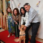 Four adults pose with a blue-vested service dog on a red carpet at a Canine Companions event