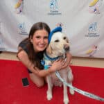 Woman kneels with a blue-vested therapy dog on a red carpet at a Canine Companions event