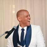 Man in a white suit and navy tie speaking at a microphone, laughing on a stage backdrop