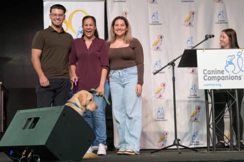 Three smiling people onstage with a yellow-vested service dog in front of a Canine Companions backdrop