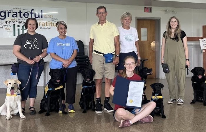 A group of seven people, some with guide dogs, posing indoors. A young girl in front is holding a certificate. The background has a wall with words like "grateful" and "appreciate" displayed.