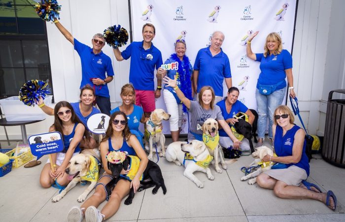 A group of people wearing blue cheer in front of a Canine Companions backdrop during DogFest North Central.