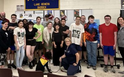 Group of people posing in a room with an FFA Lake Geneva Badger banner, featuring a seated black dog wearing a yellow and red vest.