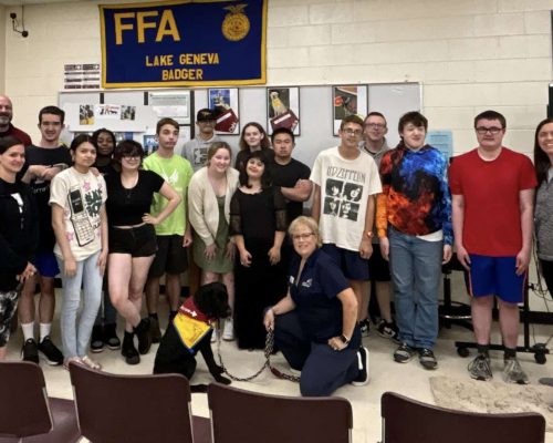 Group of people posing in a room with an FFA Lake Geneva Badger banner, featuring a seated black dog wearing a yellow and red vest.