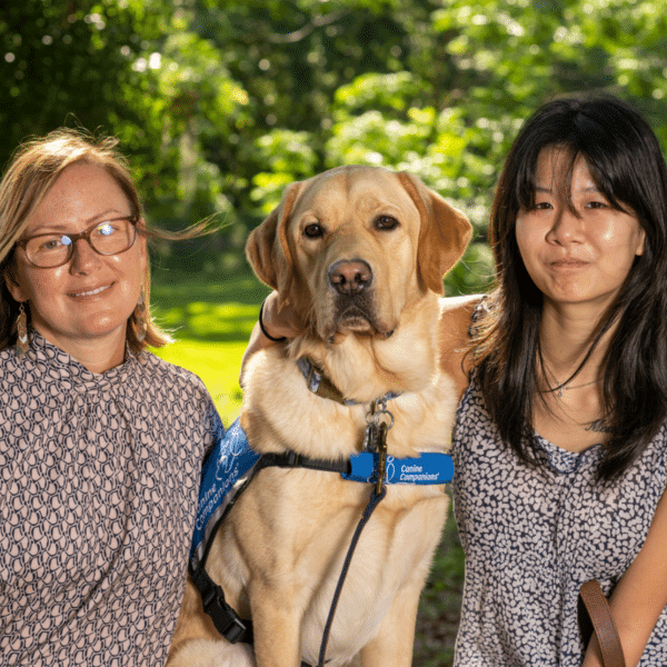 Two women pose beside a yellow Labrador Retriever wearing a blue service dog vest. The woman on the left wears glasses and a patterned blouse, while the woman on the right has long dark hair and a patterned top, both smiling warmly in a green outdoor setting.
