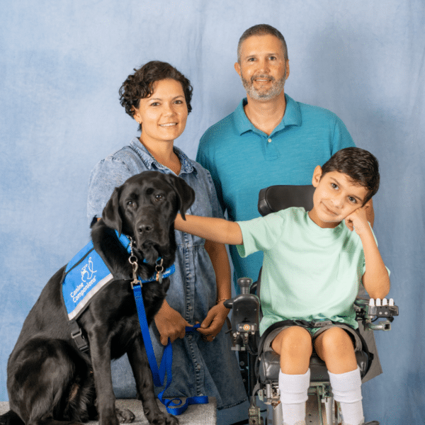 A family portrait featuring a woman and a man standing beside a boy in a wheelchair, along with a black service dog wearing a blue vest. The woman has short, curly hair and a denim dress, while the man wears a turquoise polo shirt. The boy sits in the wheelchair with his hand on his chin, wearing a light green shirt and shorts. They are set against a light blue backdrop.