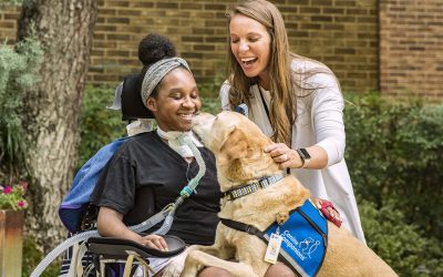 woman in a wheelchair smiling at canine companions facility dog resting on her lap while being pet by female doctor in white blouse