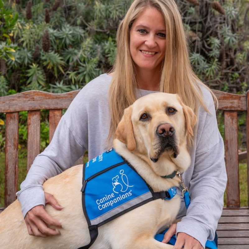 A woman with long blonde hair sits on a wooden bench, smiling and petting a yellow Labrador retriever wearing a blue harness with the logo 'Canine Companions'. The background features greenery and plants.