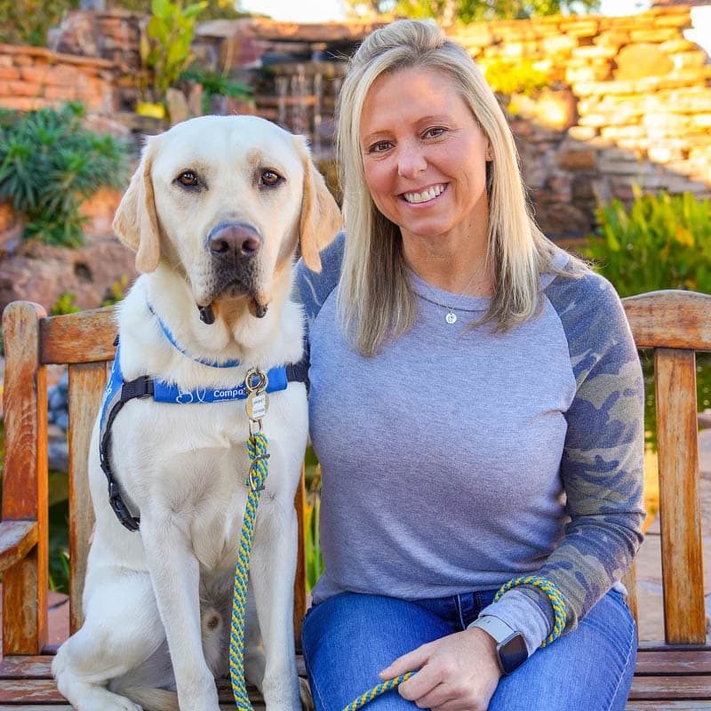 a smiling woman with a yellow labrador retriever in a blue service vest