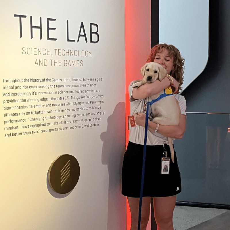 A woman with curly hair holds a Labrador puppy in her arms, smiling as she stands next to a wall exhibiting information about science and technology in sports. The wall features a section titled "THE LAB" with text discussing how innovation affects athletic performance.