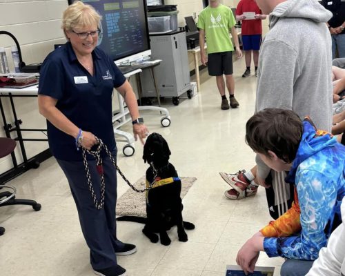 A woman demonstrating with a service dog to a group of seated students in a classroom.