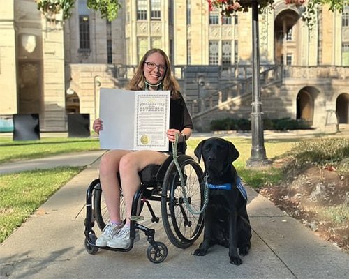 Smiling woman in a wheelchair holding a certificate sits on a campus walkway with a black service dog wearing a blue vest.