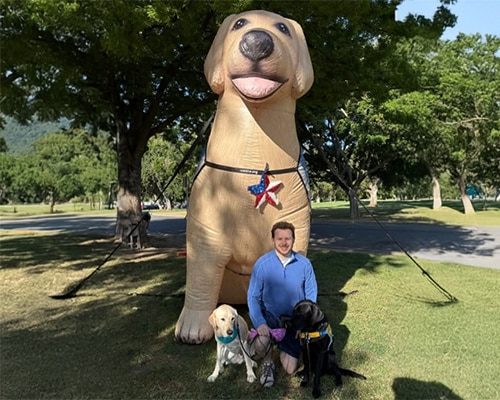 Man kneeling with three leashed dogs in front of a giant tan inflatable dog statue adorned with a star charm.