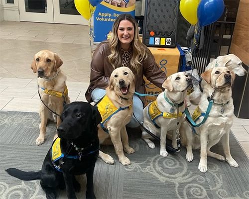 Woman kneeling and smiling with five service dogs wearing yellow vests in a lobby beside balloons and an information table.
