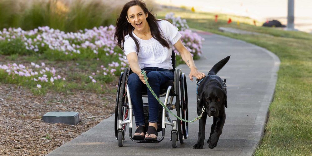 a woman in a wheelchair being pulled along a park path by her black lab service dog