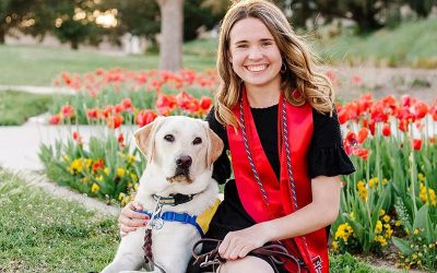 a smiling girl sitting next to a yellow lab in a yellow puppy vest